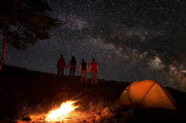 两对徒步旅行者抱着手, 在星空下, 在山上的篝火和橙色<strong>帐篷</strong>附近露营的夜晚, 在星光灿烂的天空中享受银河。浪漫的夜晚, 在神奇的星空下