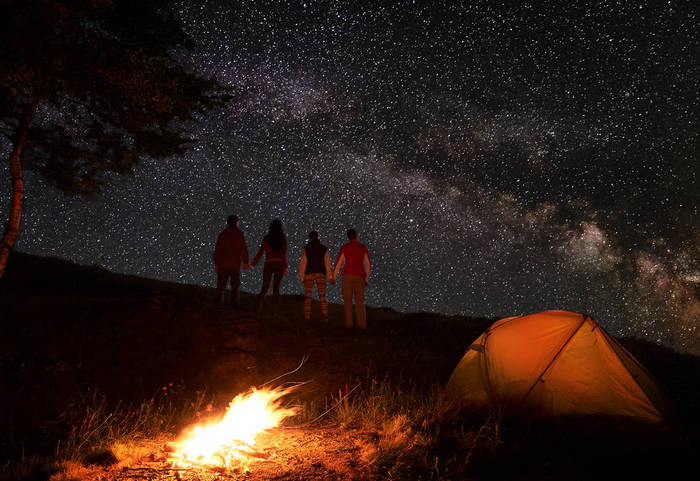 两对徒步旅行者抱着手, 在星空下, 在山上的篝火和橙色帐篷附近露营的夜晚, 在星光灿烂的天空中享受银河。浪漫的夜晚, 在神奇的星空下