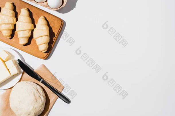 生面团、刀片、黄油和白底鸡蛋附近木制切菜板上新鲜羊角面包的顶部视图