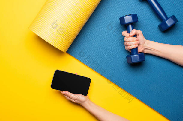 黄色背景的蓝色健身垫上拿着哑铃和智能手机的女人的剪影