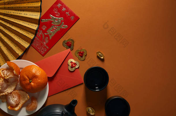 盘子上的橘子, 红茶, 带有象形文字的扇子, 金色的装饰和棕色背景上的红色信封的顶部视图