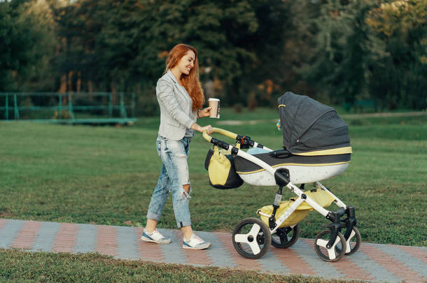 一个年轻的母亲喜欢公园散步在早上, 喝着热茶, 与她的小孩在他的婴儿推车放松.
