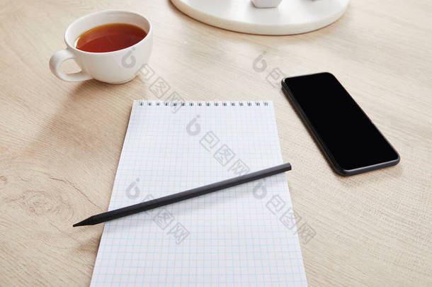 木制<strong>表</strong>面智能手机旁边的茶杯和铅笔空白笔记本