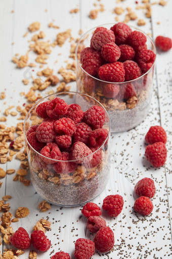 在白色木桌上散落的燕麦、浆果和种子的玻璃杯中,将树莓、燕麦片和酸奶与嘉种子放在一起图片