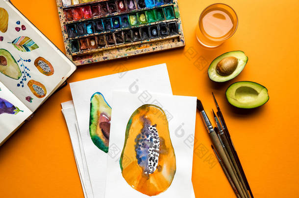 <strong>彩色</strong>油漆, 画笔, 素描本, 画与鳄梨和木瓜在黄色背景上的顶部视图