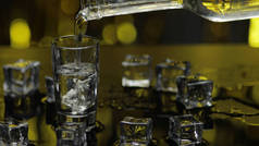 酒吧招待把冰冻伏特加从瓶子里倒进玻璃杯里.闪烁着金光的派对背景下的冰块