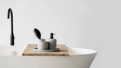 现代化的浴室内部与浴缸和水龙头。全景托盘与发刷，肥皂在瓶子分配器和干净的毛巾在木制架子上的白色当代浴室靠近黑色水龙头