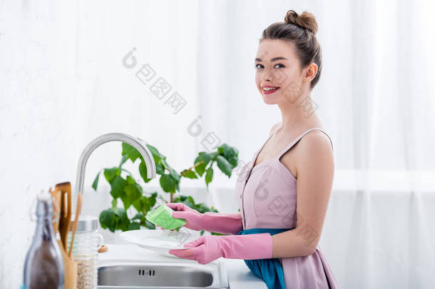 愉快的微笑的年轻妇女在橡胶手套洗涤盘在<strong>厨房</strong>