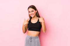 年轻的高加索健身女子穿着粉色背景的衣服，微笑着，用手指捂住嘴.