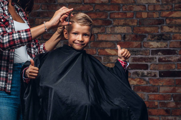 知足可爱的学龄后男孩在理发时竖起大拇指。儿童理发师与剪刀和梳子是削减小男孩在房间内阁楼内部.