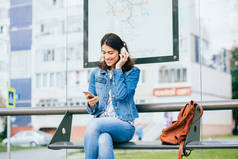 微笑兴奋的黑发女人穿着蓝色牛仔夹克与耳机望向远方，放松，听音乐，而等待在城市的电车站.