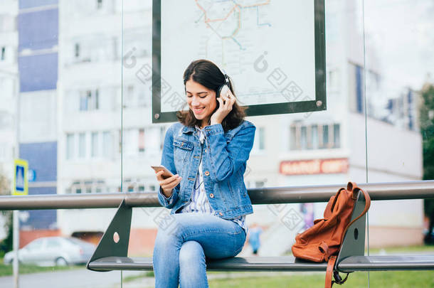 微笑兴奋的黑发女人穿着蓝色牛仔夹克与耳机望向远方，放松，听音乐，而等待在城市的电车站.