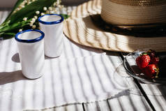 两个白瓷眼镜, 一顶帽子, 百合的山谷花束, 草莓在木桌上, 阳光洒在露台上