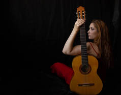 身穿红色衣服的女孩坐在靠近吉它的地板上，背景是黑色的