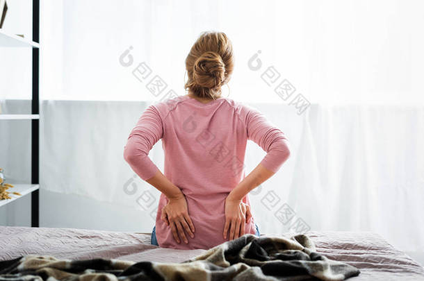 <strong>坐在</strong>公寓<strong>床上</strong>的背痛的妇女的后视图 