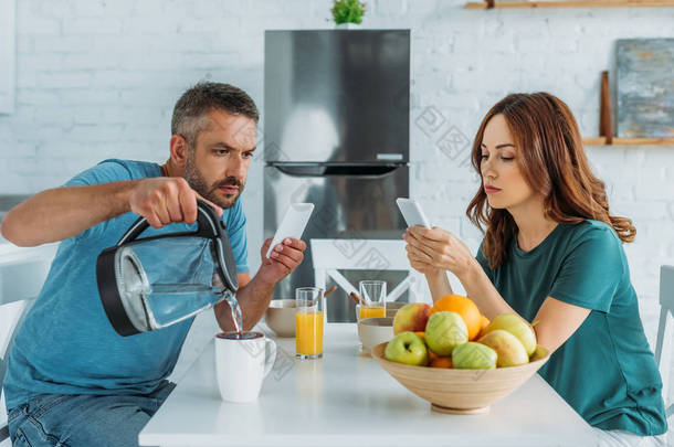 男子与智能手机<strong>倒水</strong>杯，而坐在厨房附近<strong>的</strong>妻子使用智能手机