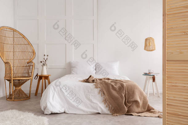 藤椅和木桌旁边的床与棕色毯子在白色卧室内饰。真实照片