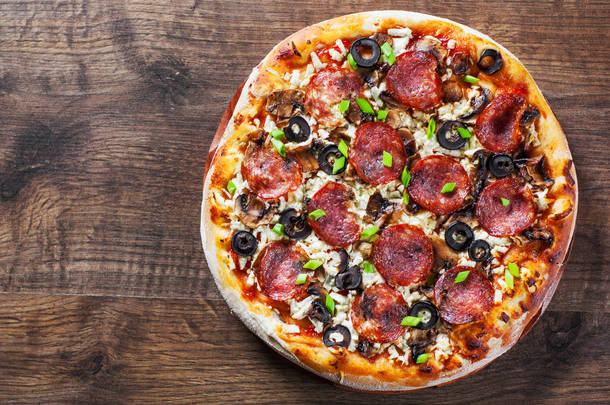 披萨配莫扎雷拉奶酪、意大利香肠、胡椒、意大利香肠、橄榄、香料和蘑菇。背景木桌上的意大利比萨饼
