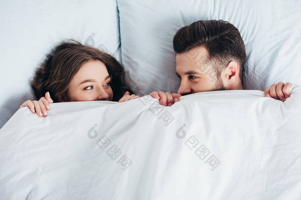 年轻的爱的夫妇躲在毯子下的床上, 看着眼睛