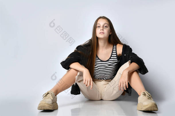 少女穿着条纹T恤、黑色斜纹棉布夹克、裤子和运动鞋。 她孤零零地坐在地板上. 靠近点