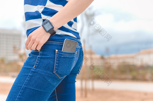 背面的牛仔裤<strong>智能</strong>手表用手机在口袋里的女人的看法。关闭.