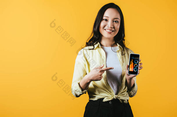 微笑的亚洲女孩指着智能手机与信息图，隔离在黄色