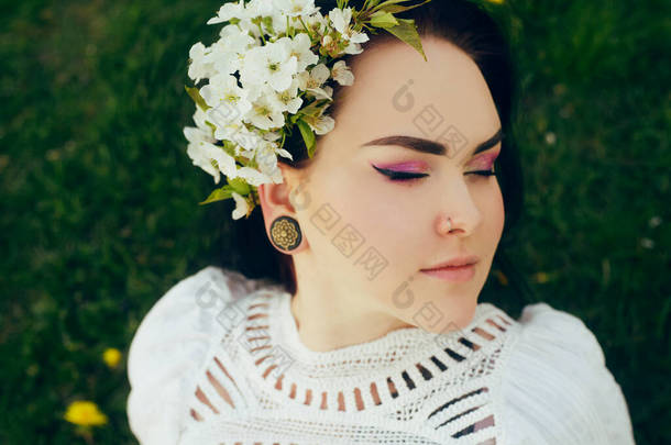那女孩躺在公园的草地上。漂亮的粉红化妆品在一个女孩身上。布鲁内特新娘穿着漂亮的布荷花裙，头发上长着花。一个穿着白色衣服的女孩的温柔照片。在大自然的公园里休息，呼吸新鲜空气.