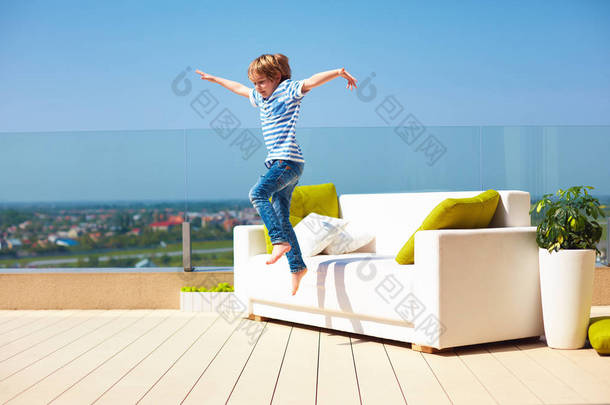 快乐的孩子，男孩从沙发上跳上屋顶顶部平台在温暖的阳光灿烂的日子