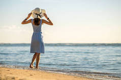 戴着草帽，身穿连衣裙的年轻女子独自站在海滨空旷的沙滩上。3.度假时,寂寞的女孩凝视着地平线,凝视着平静的海面.