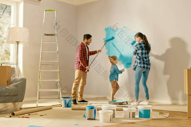 美丽的年轻<strong>家</strong>庭正在展示如何油漆墙壁,他们可爱的小女儿。他们用浅蓝色油漆覆盖的滚轮进行喷漆。房间<strong>装修</strong>在<strong>家</strong>里.