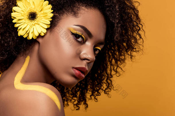 年轻嫩的非洲裔美国妇女与艺术的化妆, 在头发上寻找在橙色背景下孤立的小菊
