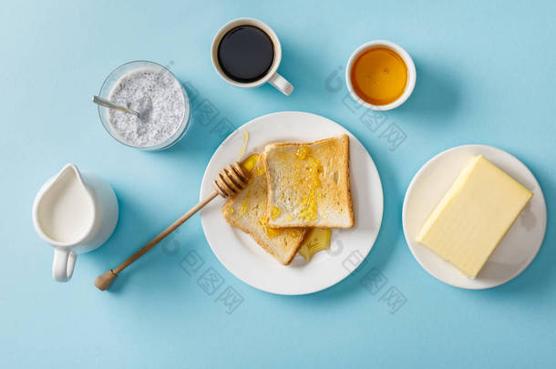 咖啡、<strong>牛奶</strong>、酸奶与奇亚种子、黄油、蜂蜜、蜂蜜烤面包和蓝色背景的白色盘子上的木制浸渍