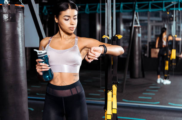 神奇的运动女孩拿着水瓶, 同时看着健身跟踪器在体育健身房 