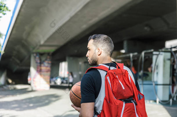 一个身强体壮的篮球运动员背着一个大的红色背包，一边走路一边打篮球的背影