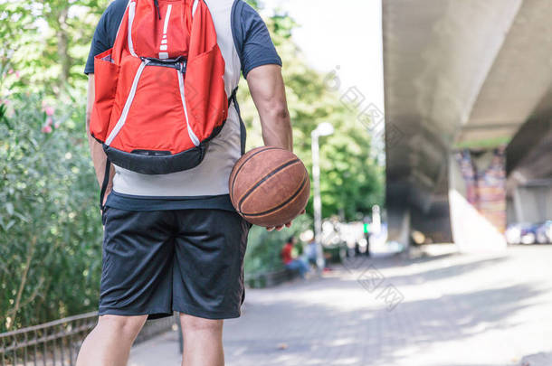 一个男篮球员背着他的球和一个红色的大背包在公园里散步的背影