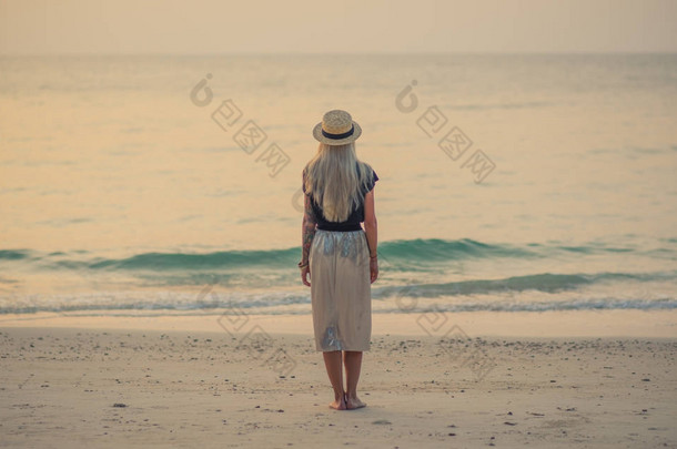 年轻的金发女郎站在清晨的海面上, 看着平静的绿松石水。清晨在海边漫步。站在他的背部相机.
