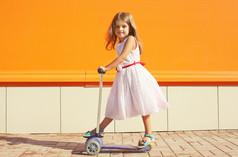 在反对多彩墙坝滑板车上的衣服的小女孩