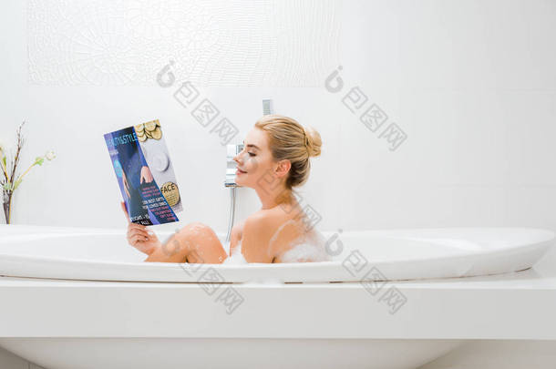 有吸引力的和金发碧眼的女人洗澡与泡沫和阅读美容和风格杂志在浴室的侧视图 