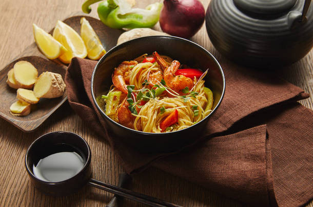 在筷子、酱油、柠檬和生姜根、餐巾上的茶壶旁边的碗里放着虾仁和蔬菜的面条