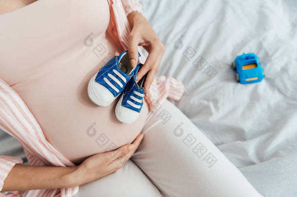 孕妇与玩具车和婴儿鞋坐在床上和触摸腹部的部分视图