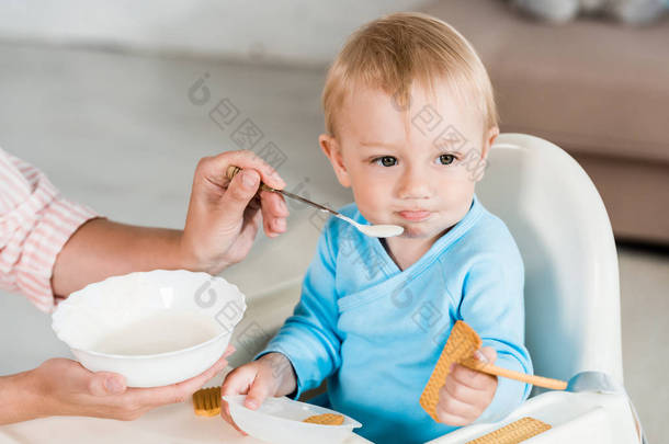 裁剪视图的母亲拿着碗和喂养可爱的幼儿儿子在家里 