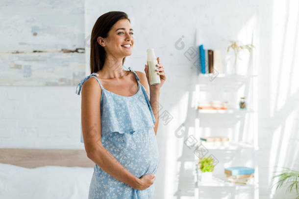 穿着蓝色衣服的快乐孕妇在卧室里拿着一瓶牛奶 