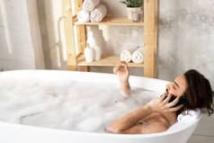 年轻而安静的男人一边躺在浴缸里一边用智能手机说话，一边在浴室里满满的水和泡沫