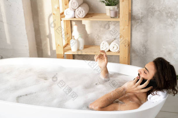 年轻而安静的男人一边躺在浴缸里一边用智能手机说话，一边在浴室里满满的水和泡沫