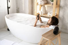 年轻而安详的男人躺在浴缸里，在浴室的角落里装满水和泡沫，一边用智能手机