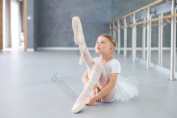 芭蕾班里的小芭蕾舞演员正在试穿尖皮鞋. 可爱的小女孩坐在理发店下面的地板上。 那孩子穿着白色芭蕾服装，穿着迷你裙跳舞.