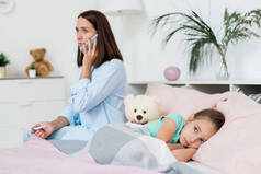 生病的小女孩躺在床上躺在毯子下，而她的母亲用手机打电话给医生