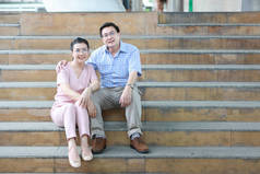 亚洲旅客夫妇夫妇在粉红色和蓝色衬衫与白色漂亮的帽子坐在市中心的楼梯上，休息与快乐的笑脸，同时拥抱 
