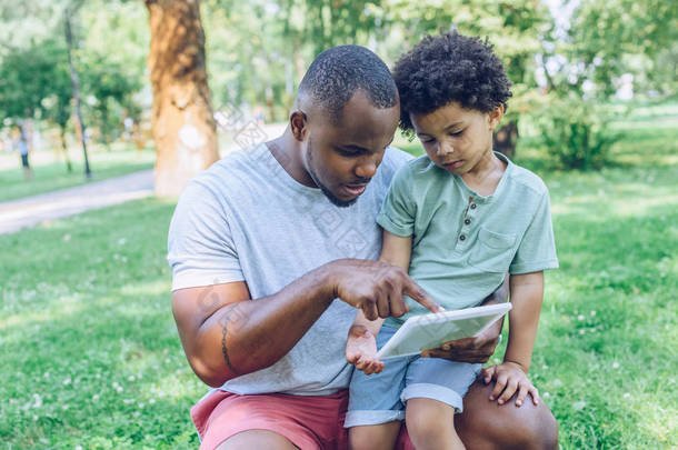 年轻的非洲裔美国父亲在公园里用数码平板电脑给可爱的儿子  