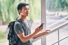 年轻的亚洲男学生手拿智能手机，穿着T恤和背包站在大学楼梯上，对未来持乐观态度